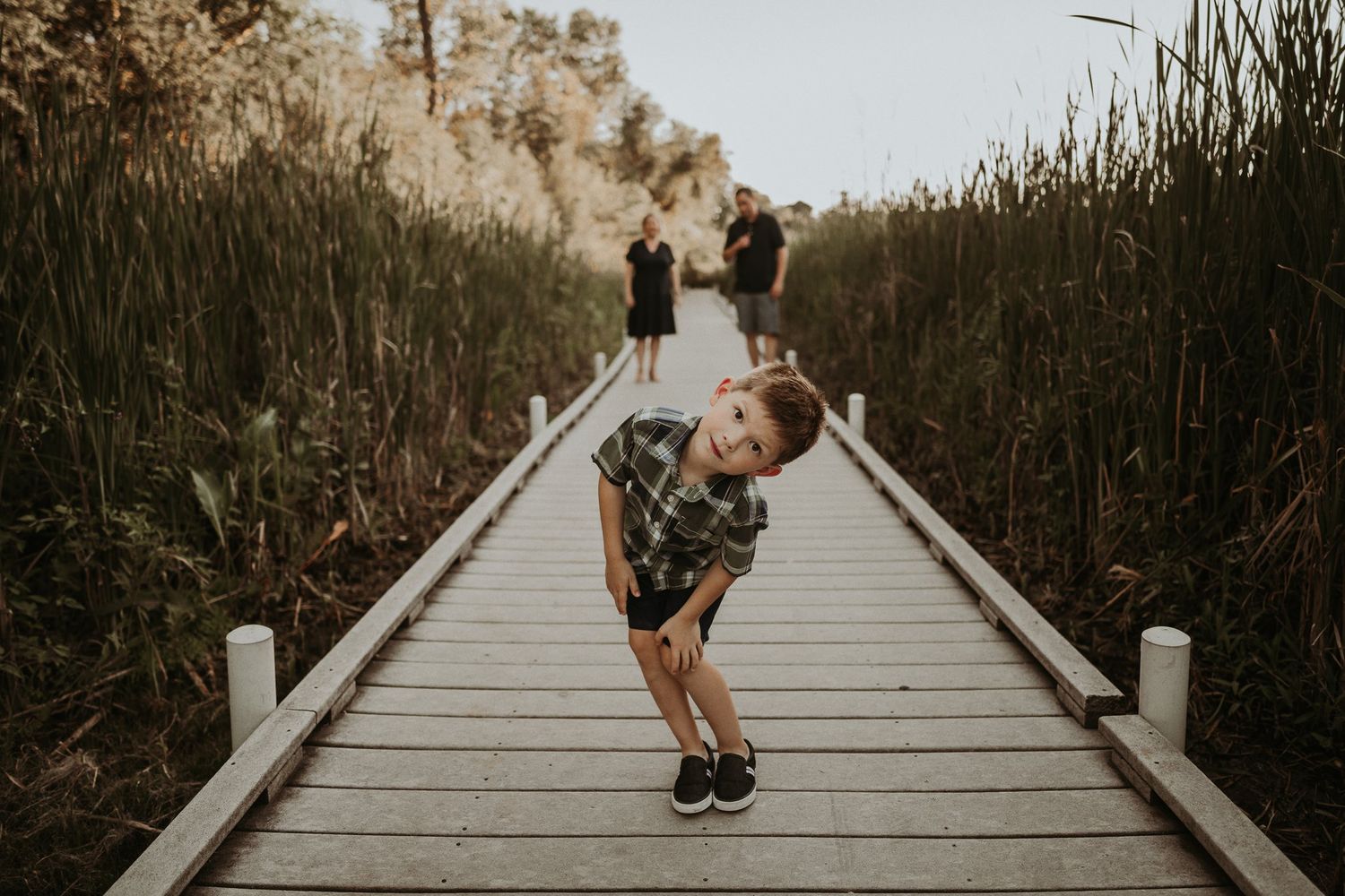 Child striking playful pose on wooden boardwalk with tall reeds in background.