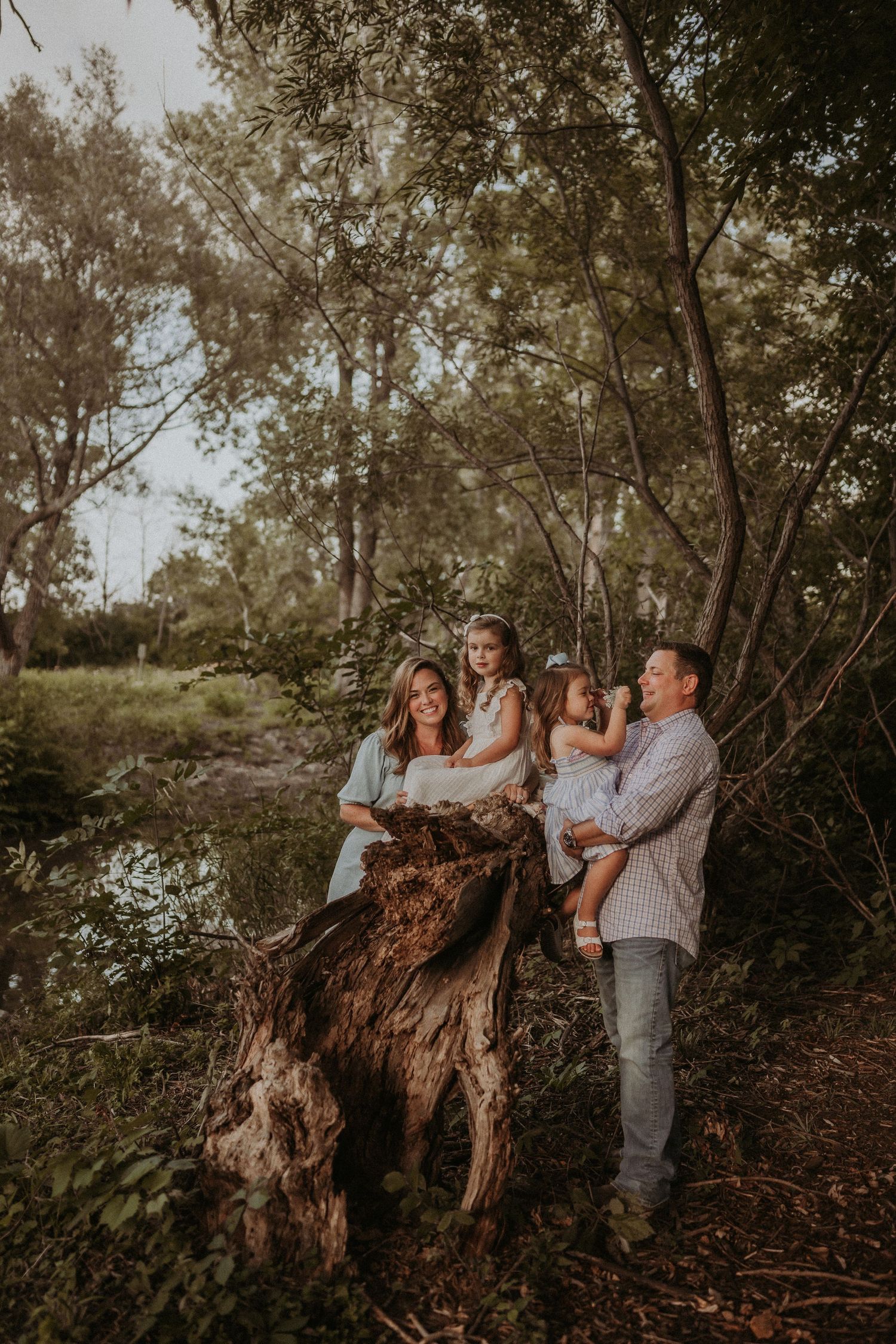 Candid family portrait by a fallen tree trunk in a dreamy woodland setting at sunset.