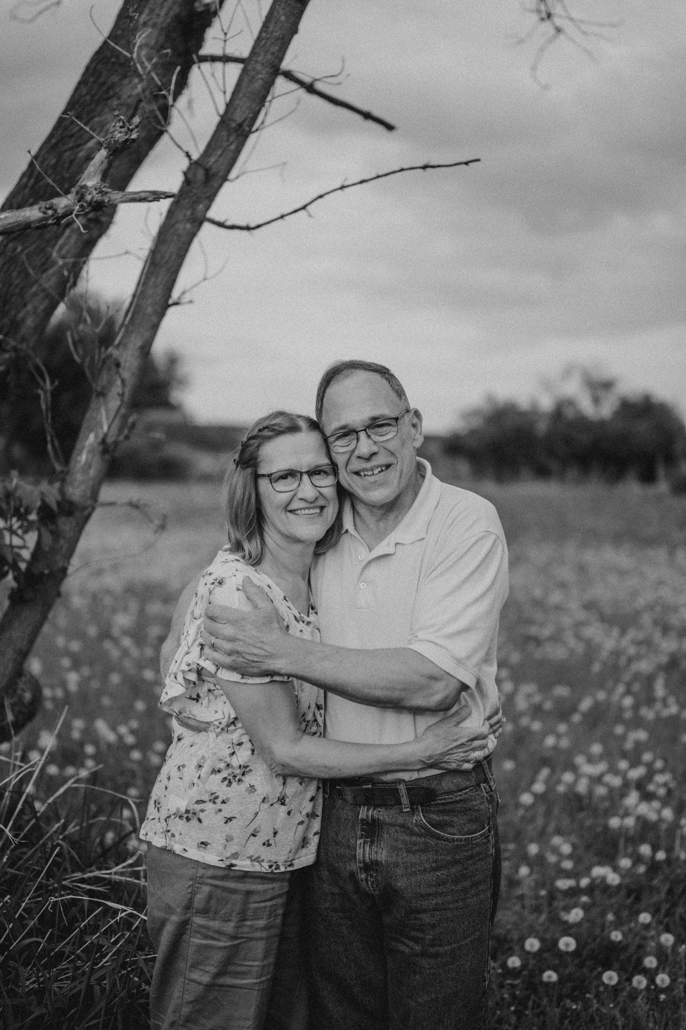 A loving couple embraces in a wildflower field on a cloudy day, captured in black and white.