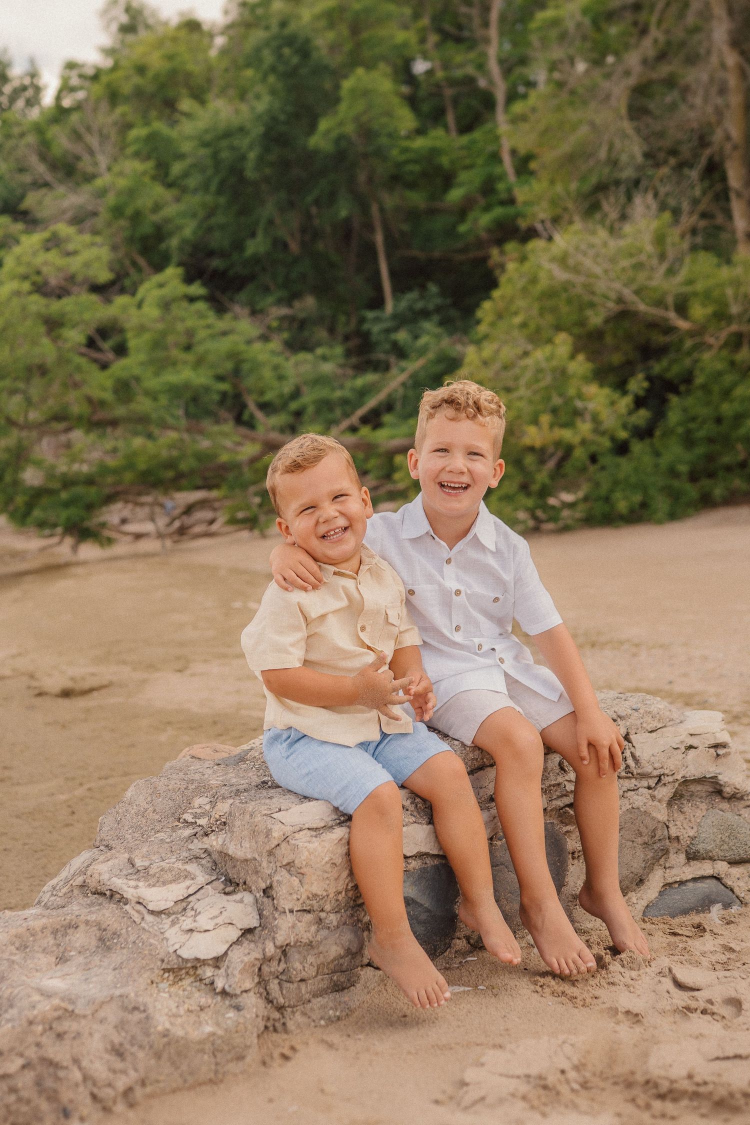 Two young children sitting together and smiling on a rocky outcrop at the beach with lush forest in background.