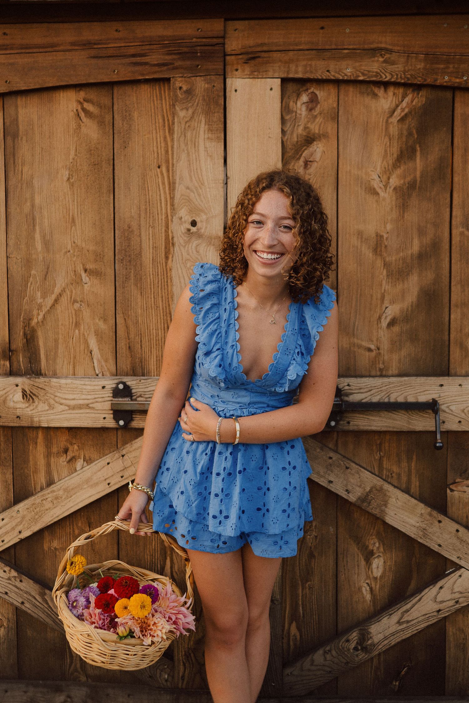 Blue romper-clad figure poses playfully in front of rustic wooden barn doors with a flower basket.