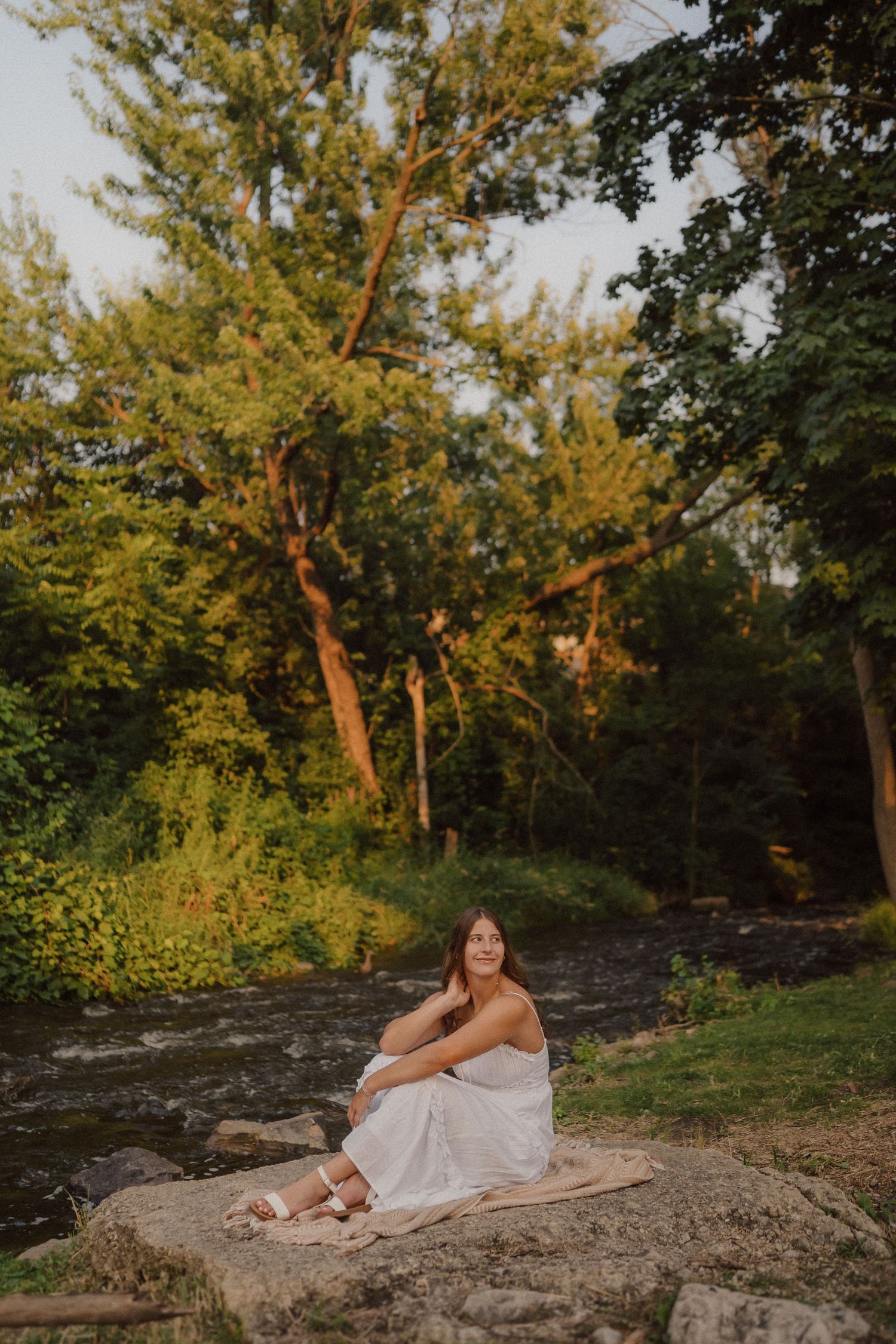 A person in white dress sits on a large rock in a natural setting during sunset.