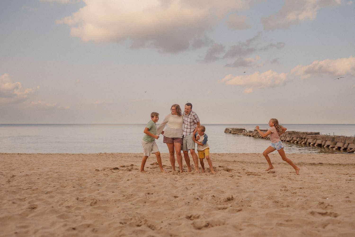 Family playing and running together on a sandy beach at sunset with a pier in the background.