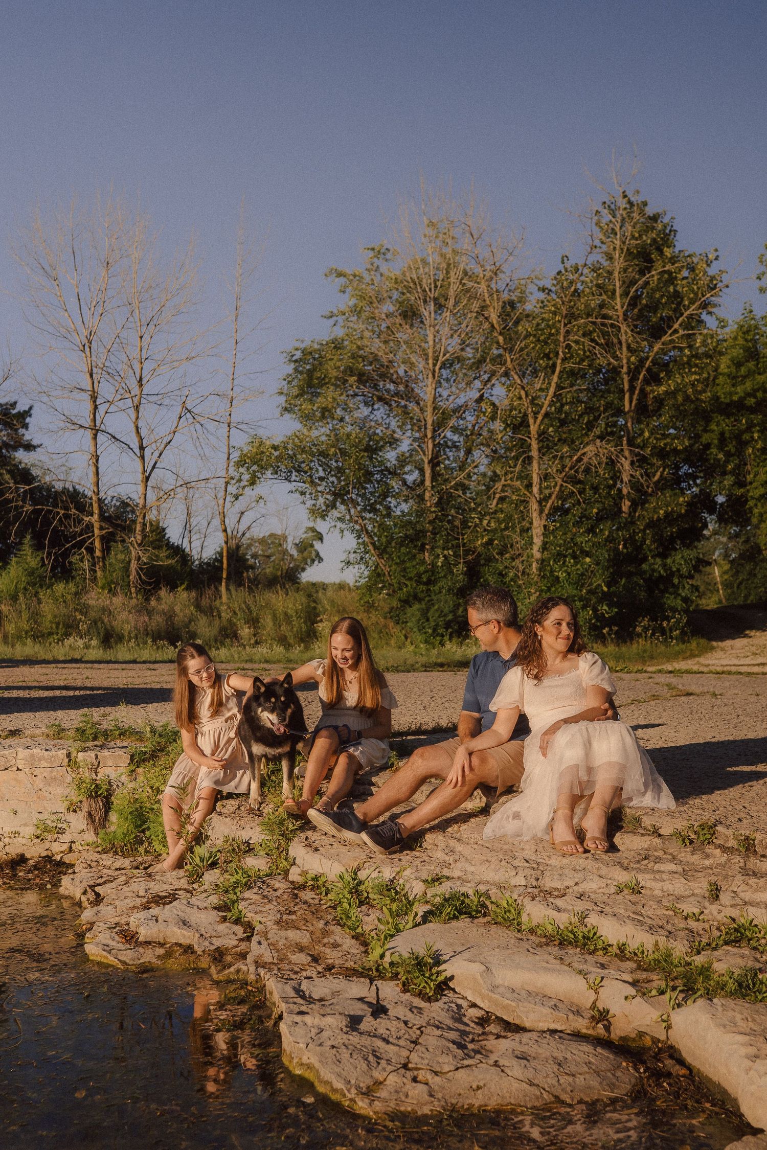 Group sitting together on a rocky riverbank at sunset with two dogs beside them.
