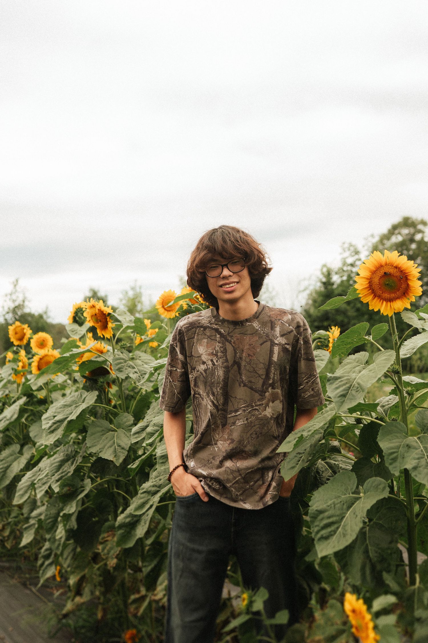 A figure in a camouflage shirt stands among tall blooming sunflowers on an overcast day.