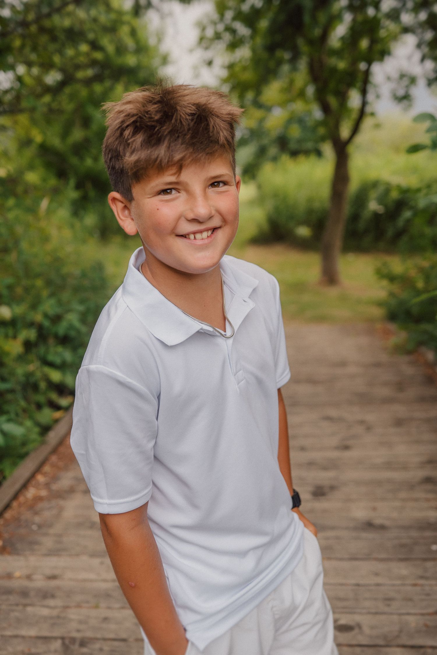 A person in a white polo shirt poses playfully on a wooden walkway surrounded by nature.