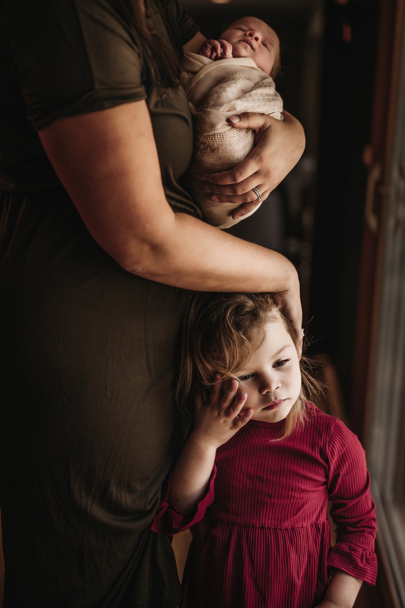Intimate family moment showing a baby wrapped in beige blanket and toddler in red dress in moody lighting.