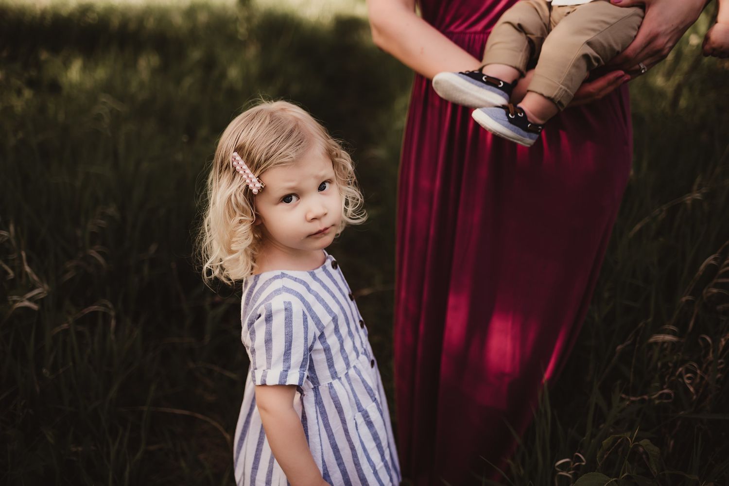 A young child in a striped dress stands in a field at sunset while someone in burgundy holds a baby.