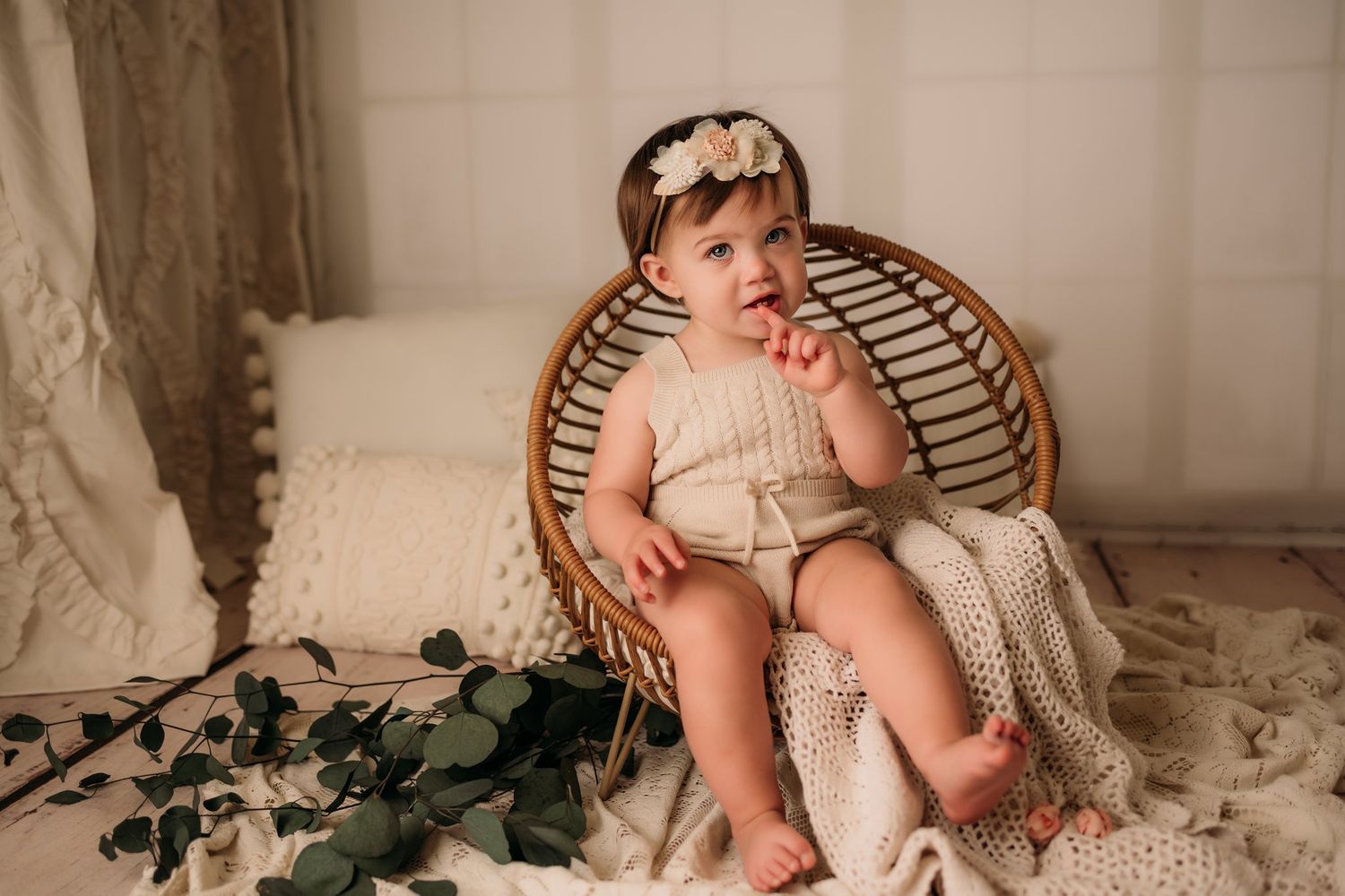 Baby sits in rattan chair with knit blanket and eucalyptus leaves as props.