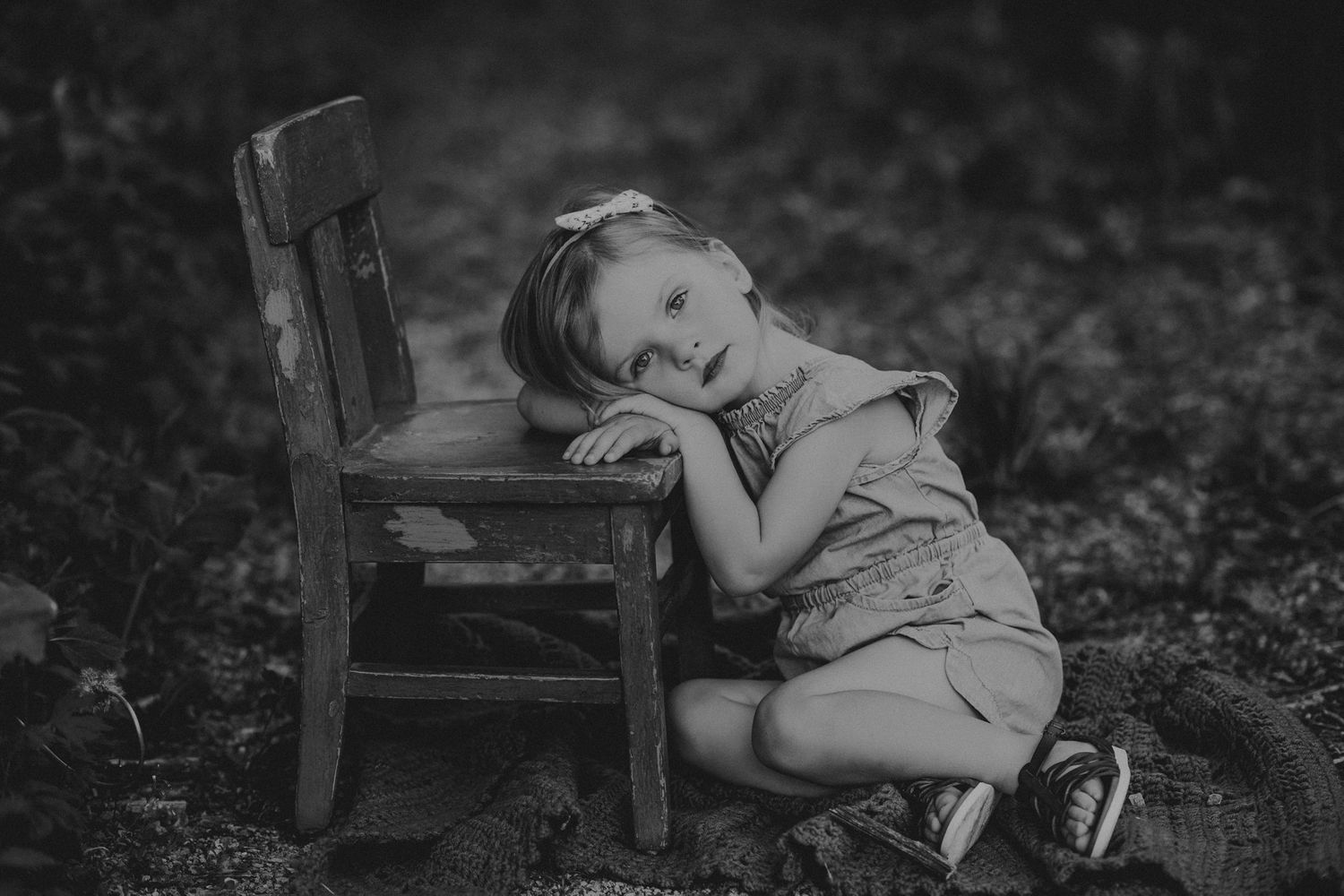 Monochrome image of child resting against rustic wooden chair outdoors.