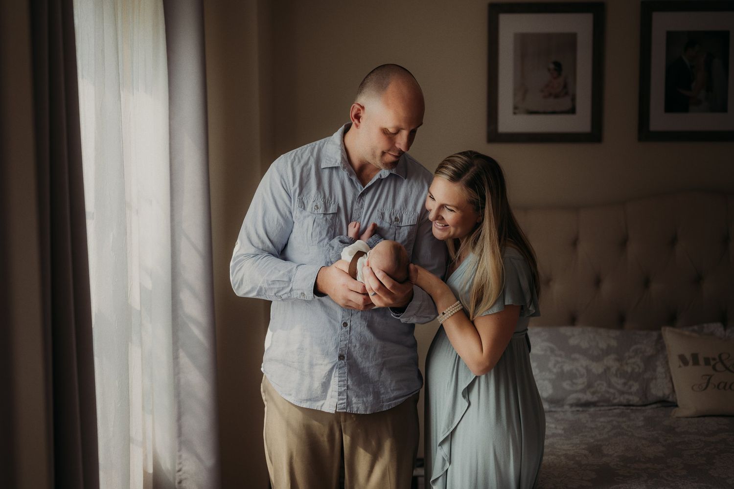 Parents share intimate moment with newborn baby near bright window.
