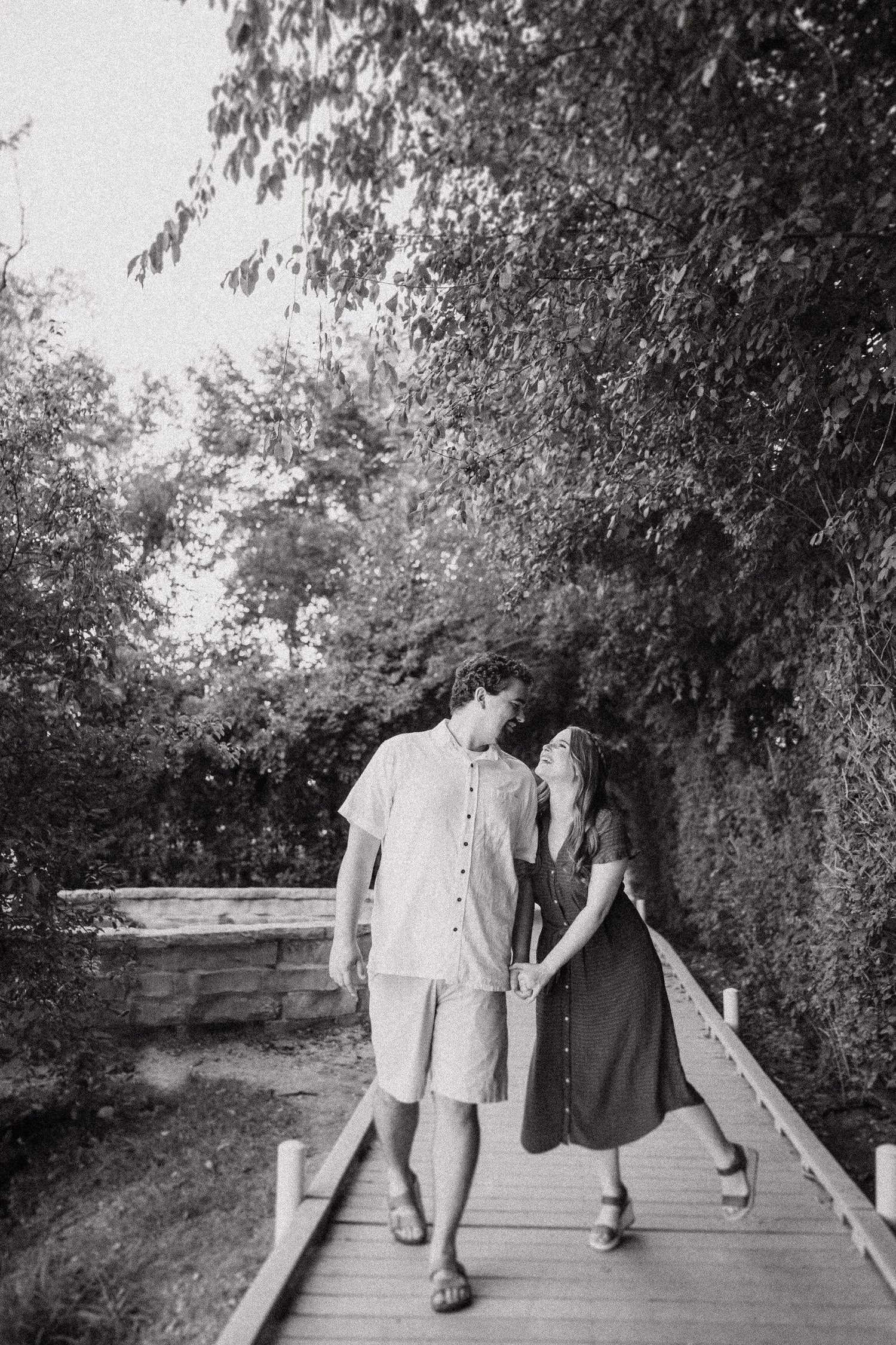 Black and white photo of two people walking together on a wooden boardwalk path through nature.