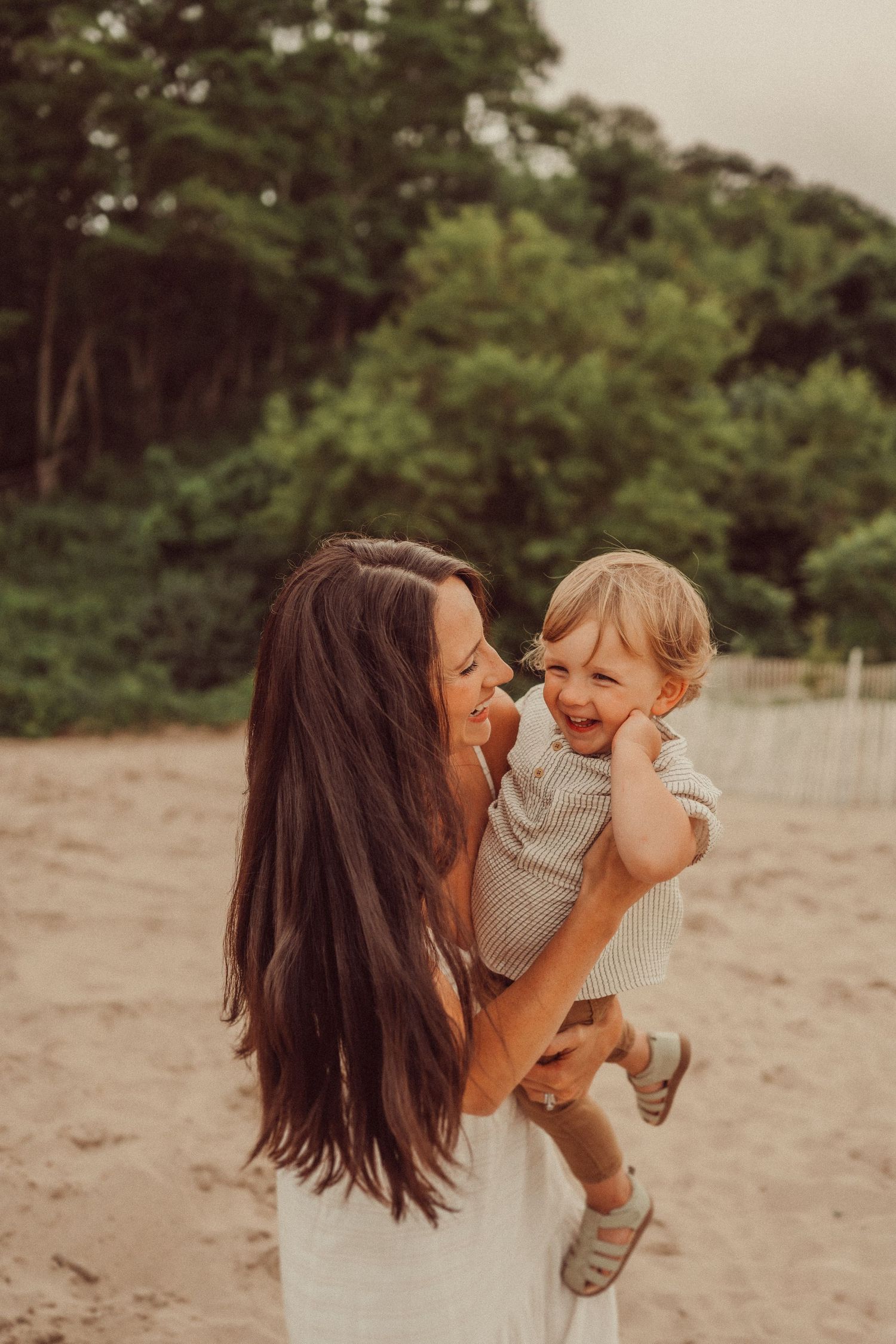 Mother in white dress holds and kisses joyful toddler during sunset beach photoshoot.