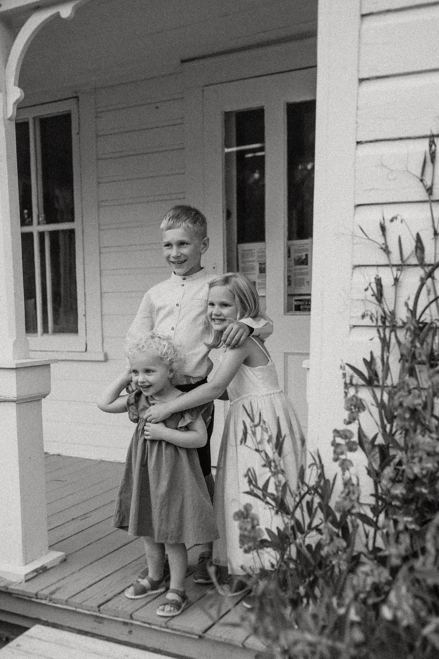Black and white portrait of siblings standing on a rustic farmhouse porch.