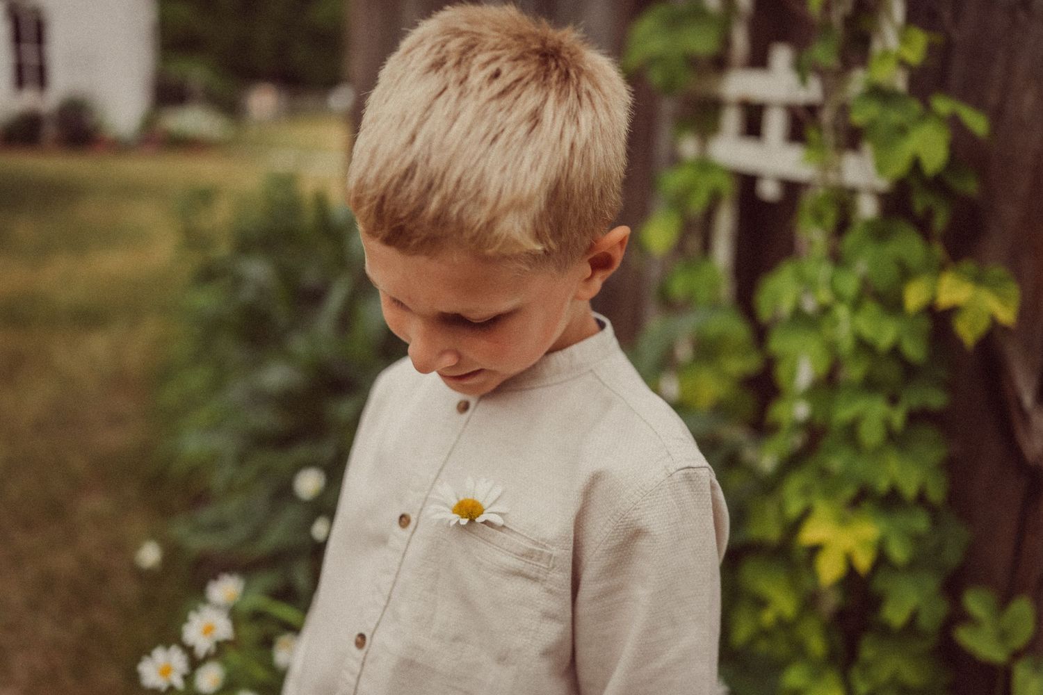 A child in a beige button-down shirt looks down while standing in a garden with climbing vines.
