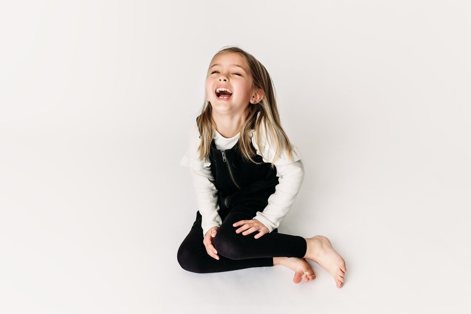 A child laughs joyfully while sitting on white background in black and white outfit.