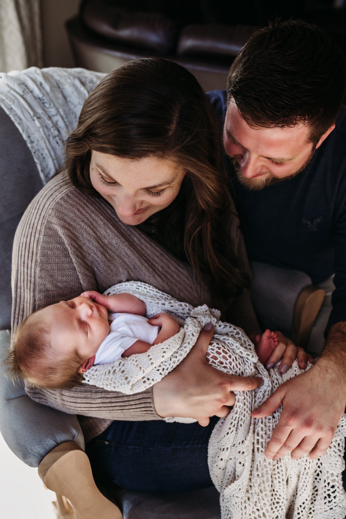 Cozy family moment on grey couch with newborn wrapped in cream lace blanket.