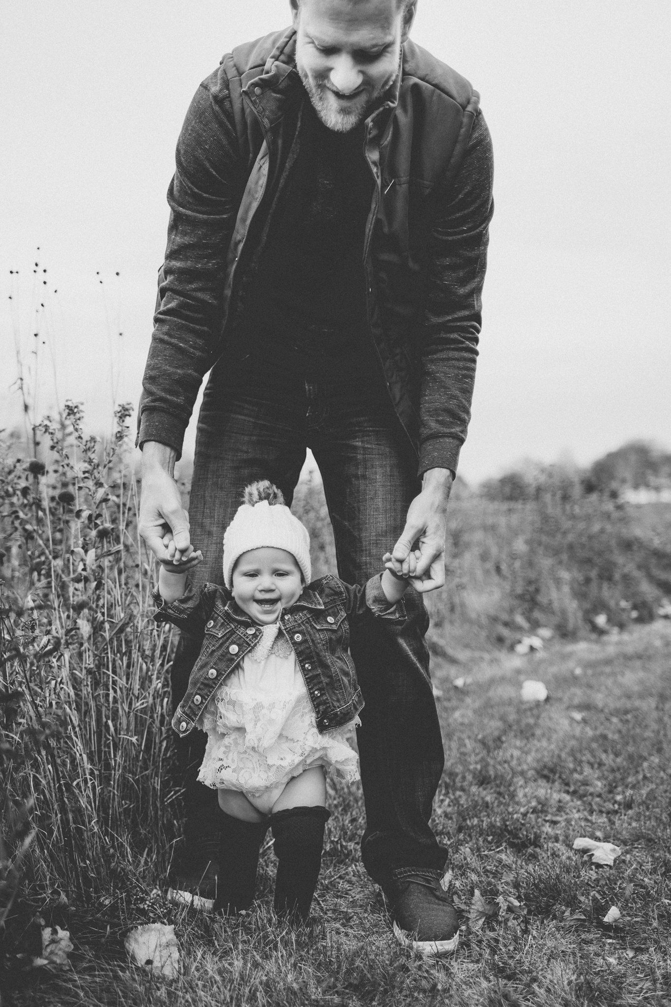 Black and white image of parent helping toddler walk through field of tall grass.