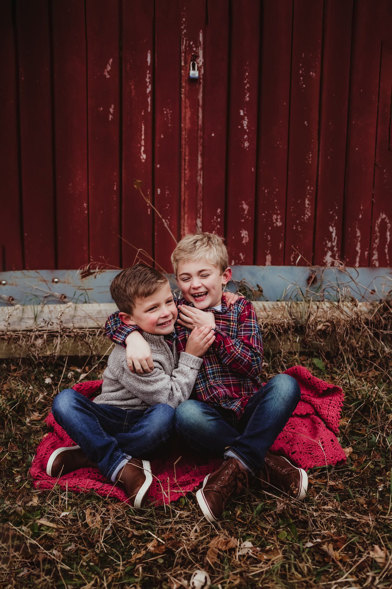 Two children share a joyful laugh while sitting on a red blanket against a barn wall.