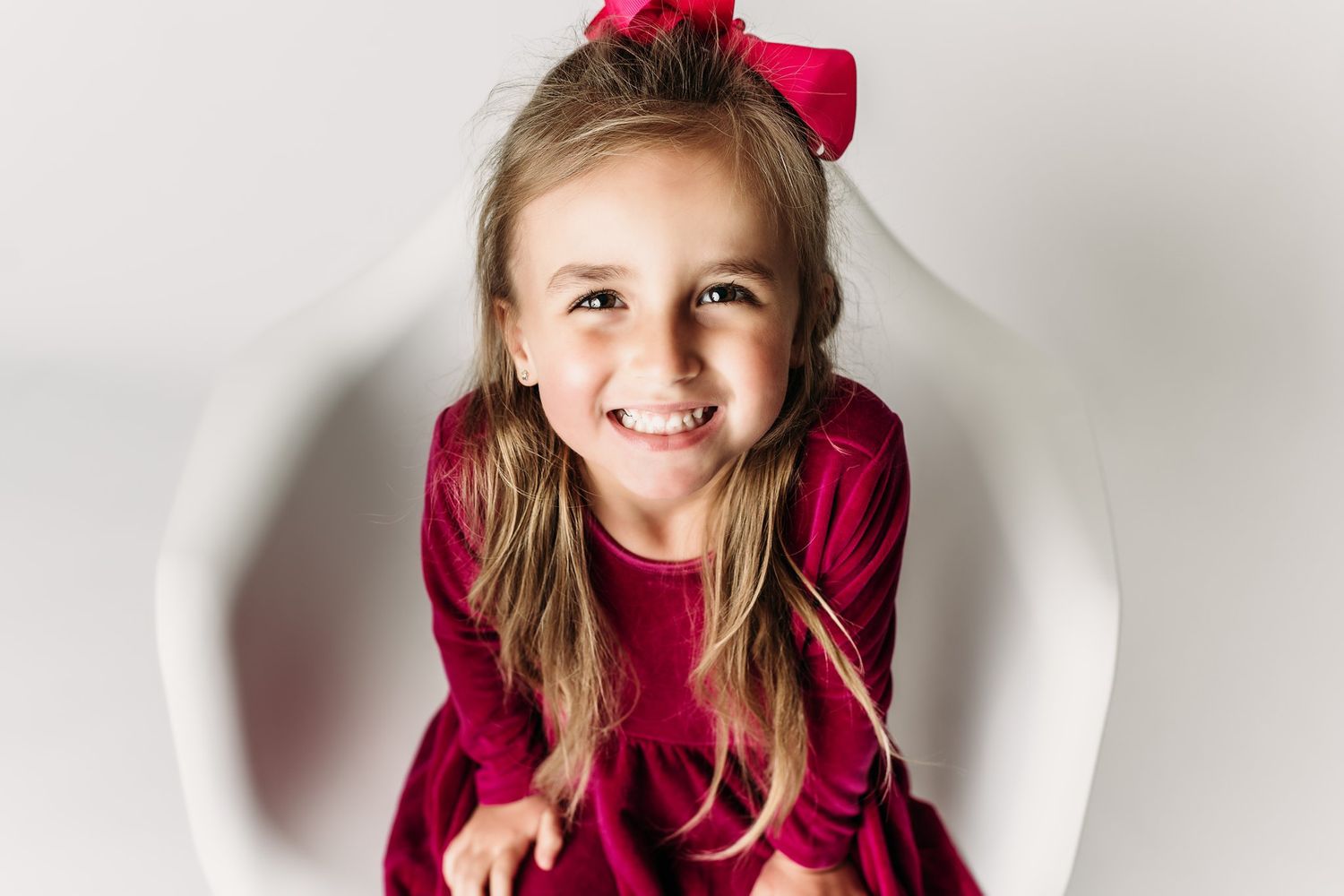 Child in velvet dress with pink bow smiling brightly on white chair.