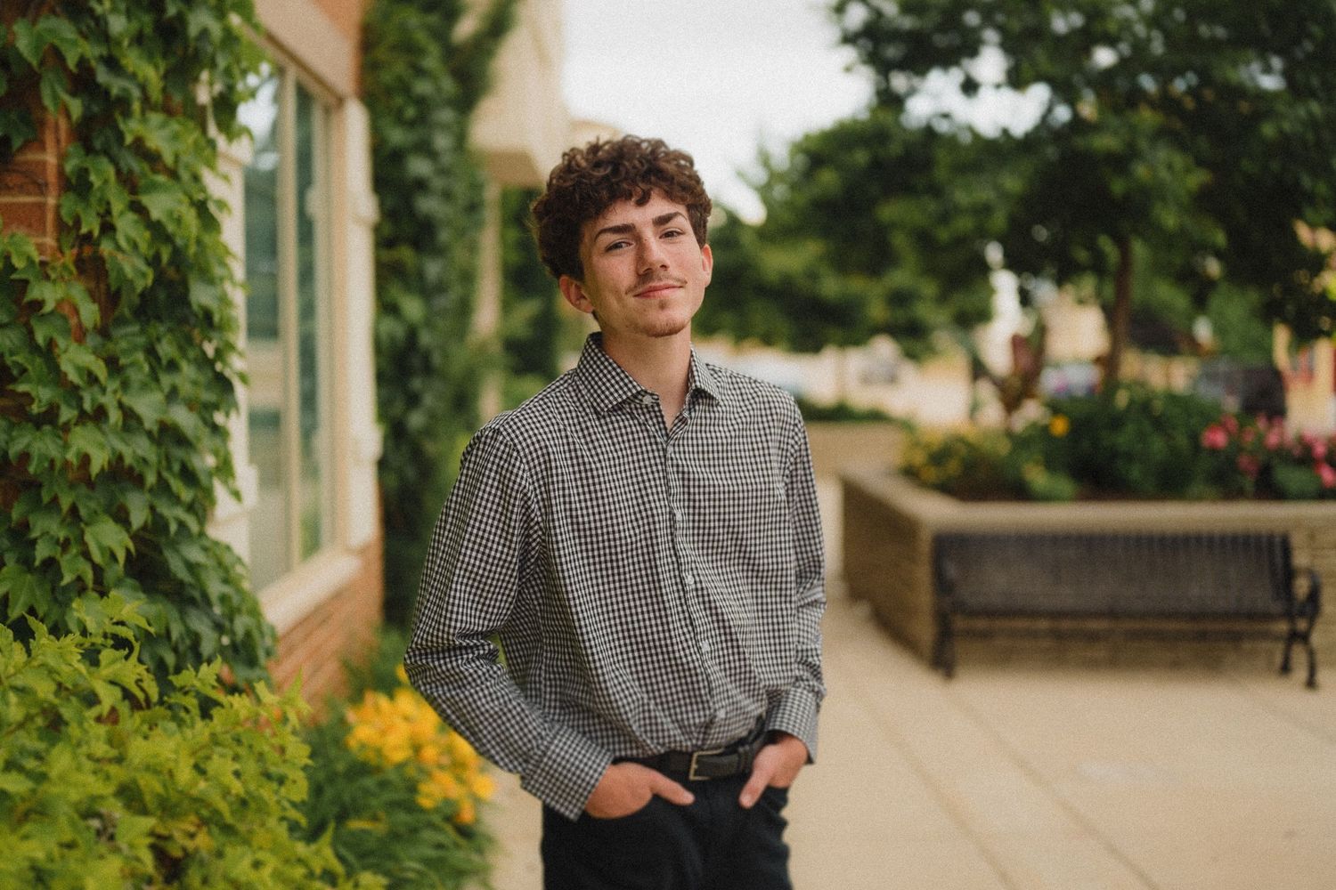 Young person in checkered button-down shirt posing against green foliage and outdoor patio furniture setting.