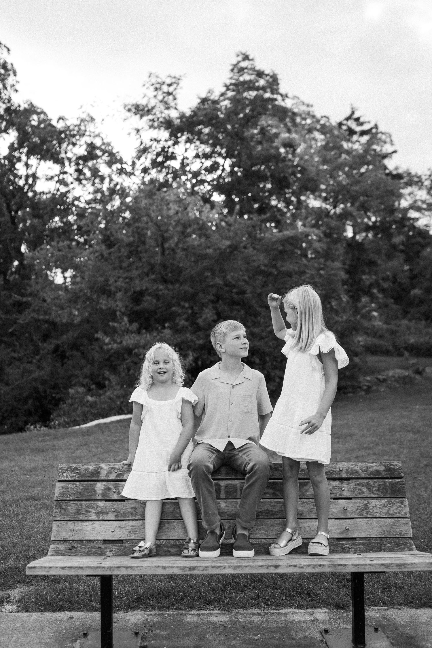 Black and white photo of people sitting together on a wooden park bench surrounded by trees.