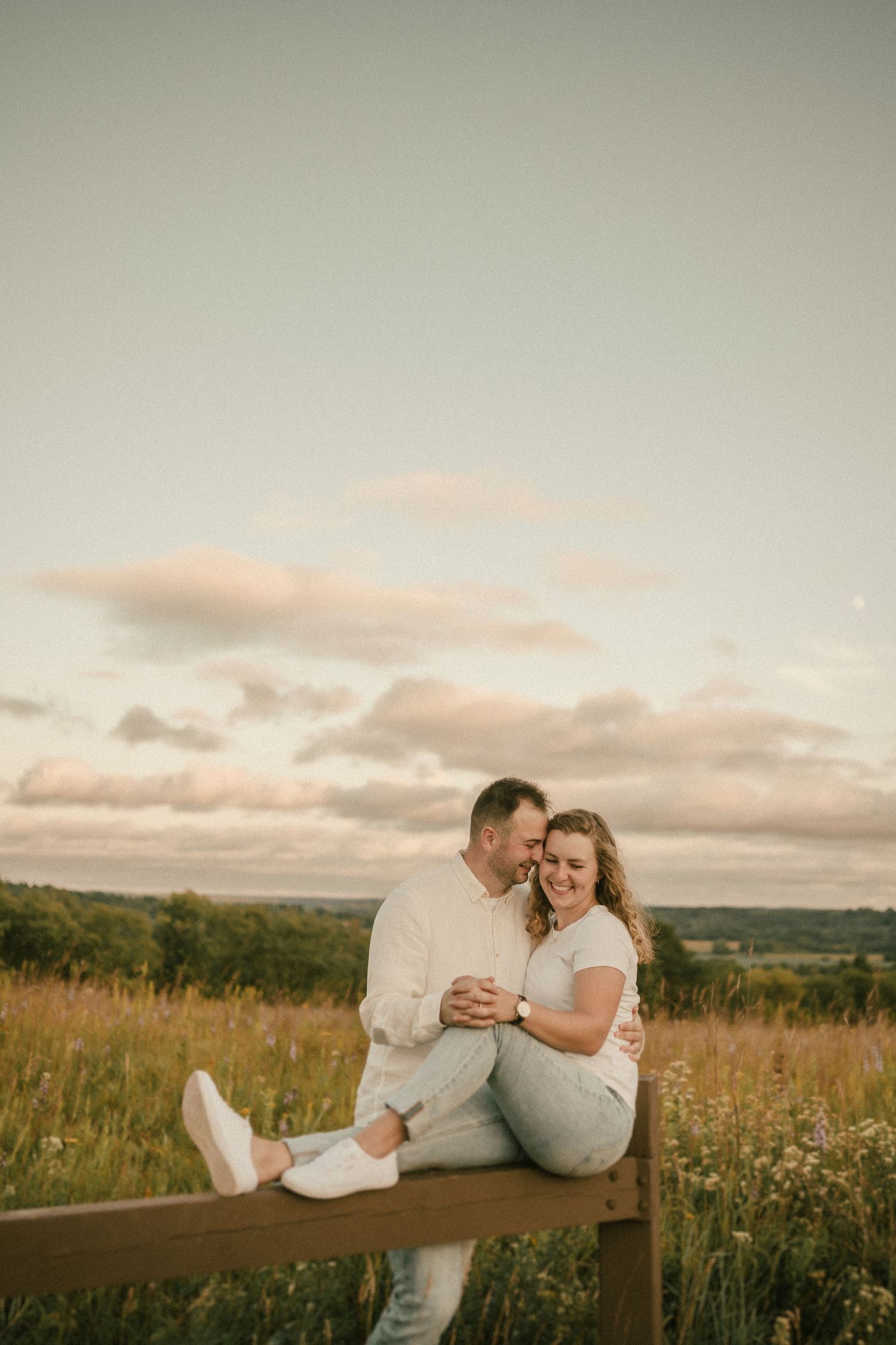 Couple sits together on wooden bench in golden meadow during sunset with rolling hills in background.