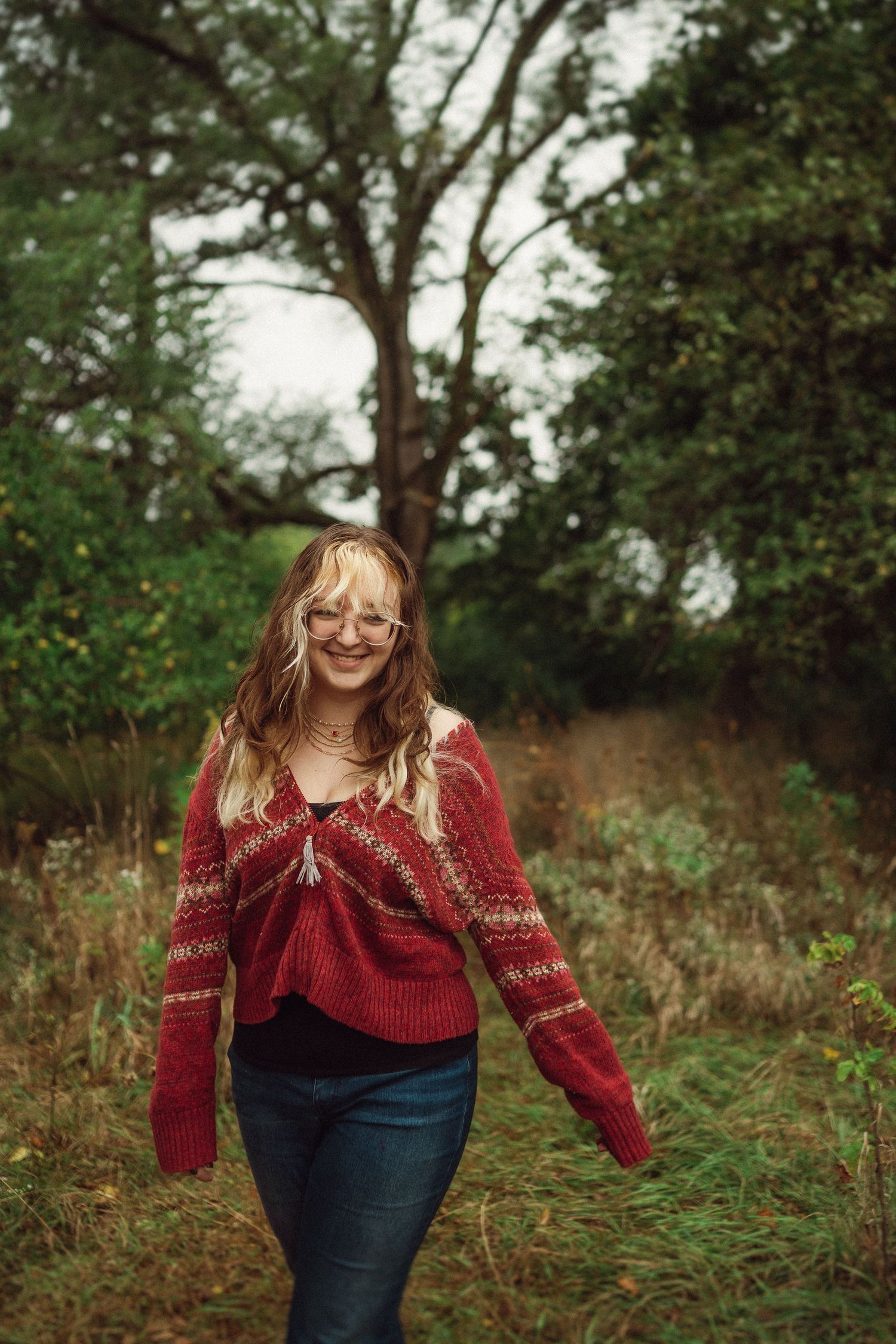 Candid outdoor photo of someone wearing a patterned red sweater and glasses in a natural setting.