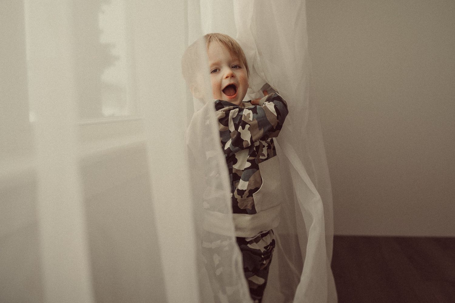 Small child playing peek-a-boo between white curtains in patterned pajamas.