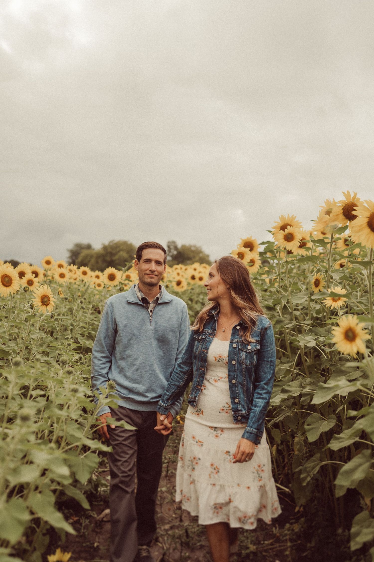 A couple walks hand in hand through a blooming sunflower field under cloudy skies.