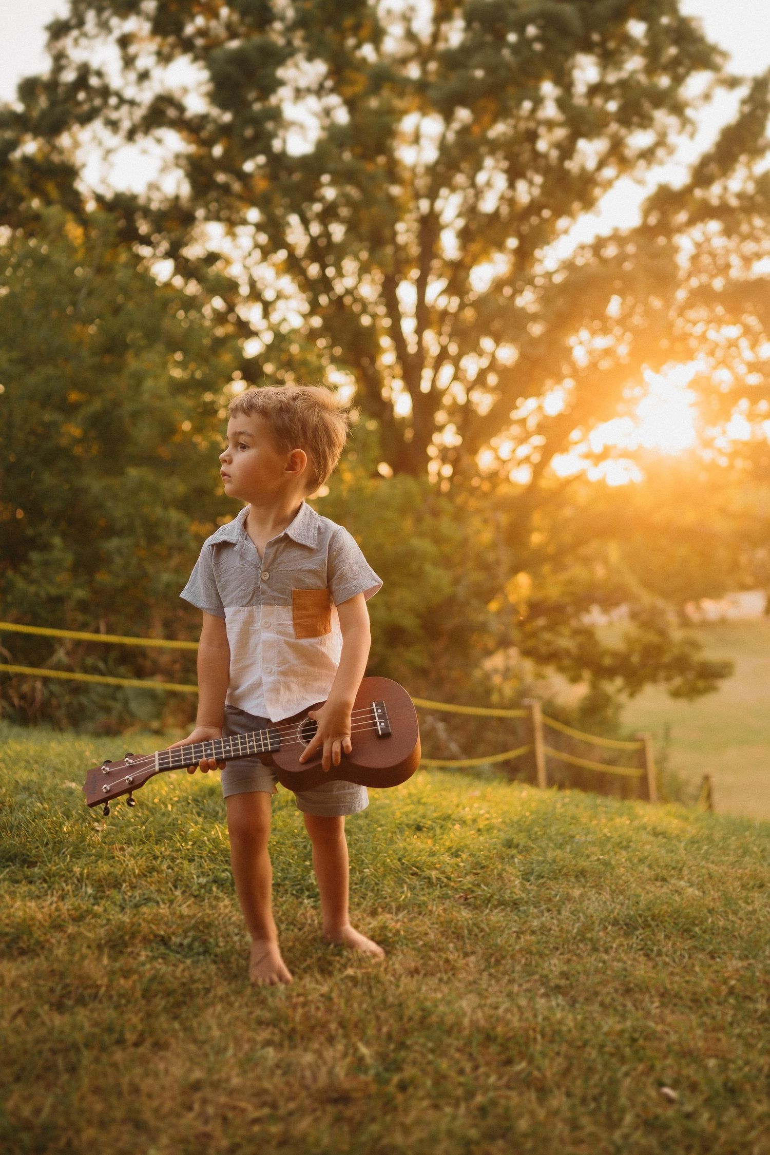 Child holding a ukulele while playing barefoot in a sunlit garden setting.
