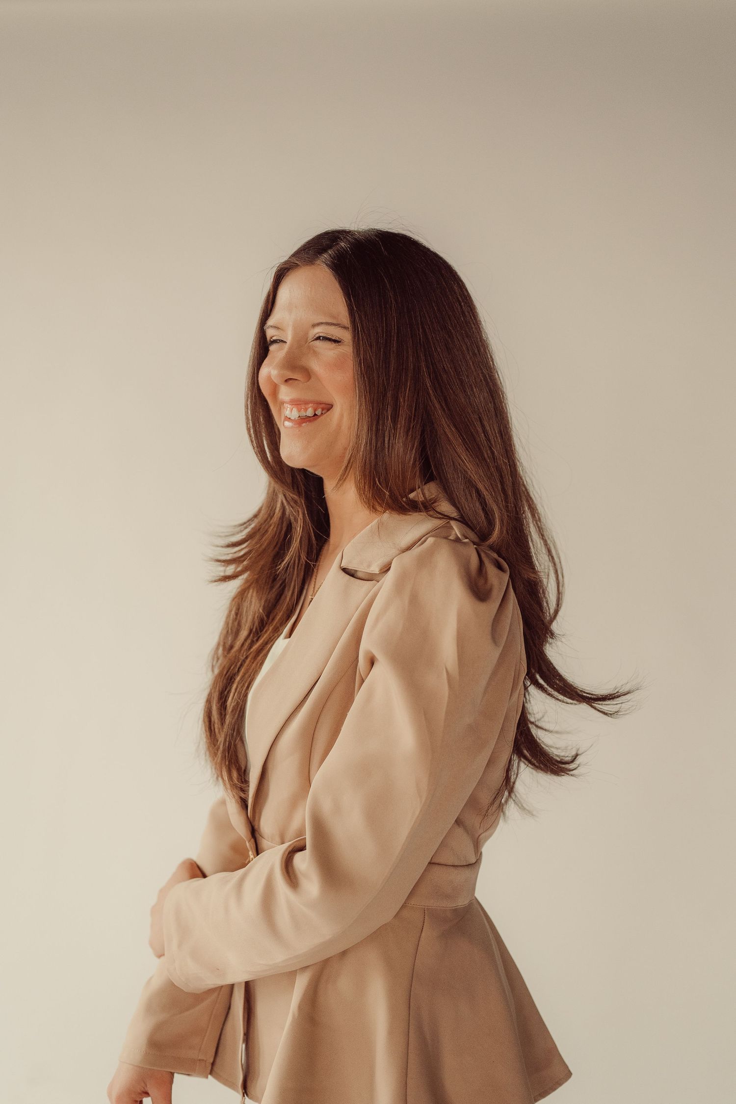 Candid profile shot of woman in beige blazer laughing against neutral backdrop.