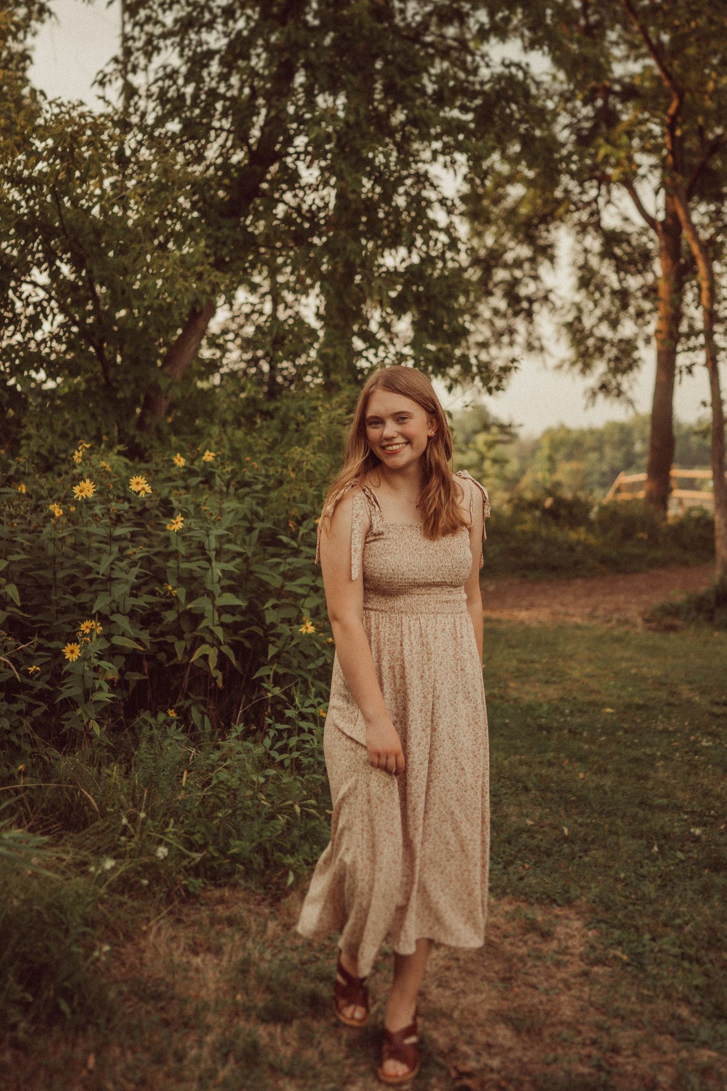 Warm toned portrait in a beige dress standing on a dirt path surrounded by sunflowers and trees.