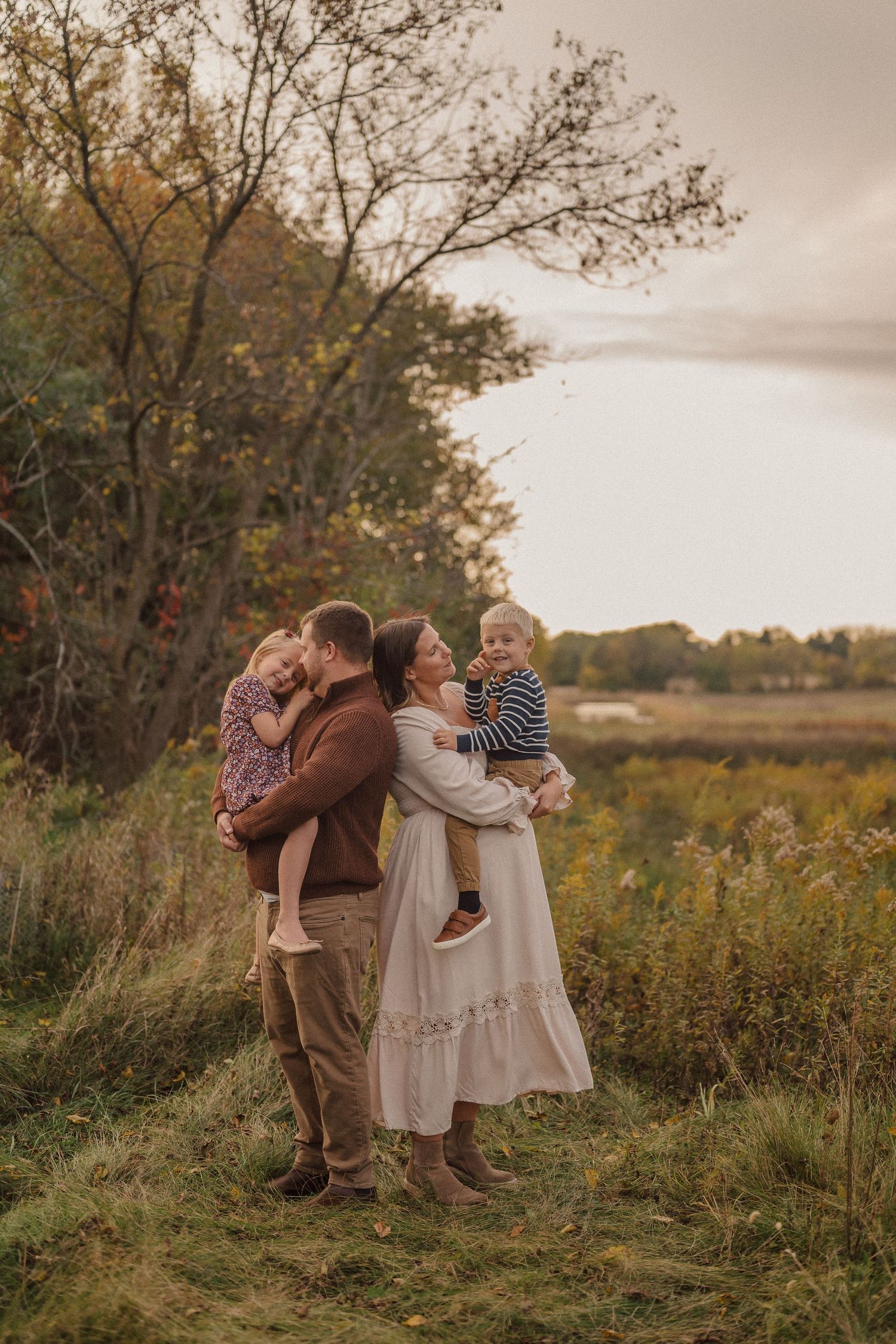 A tender family moment captured outdoors in a natural setting with tall grass and trees at sunset.