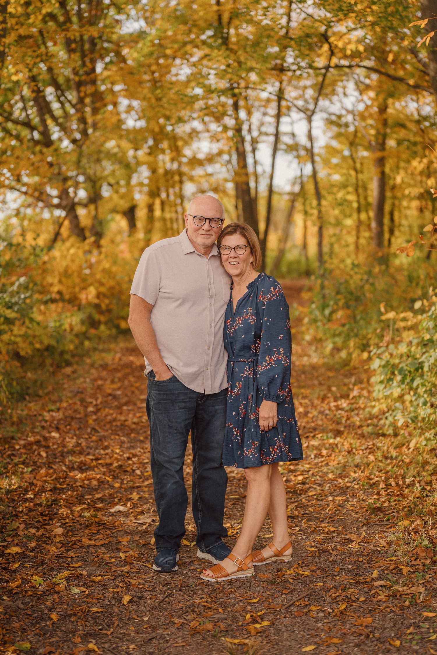 A couple shares a romantic moment on a leaf-covered path surrounded by golden autumn trees in warm sunlight.