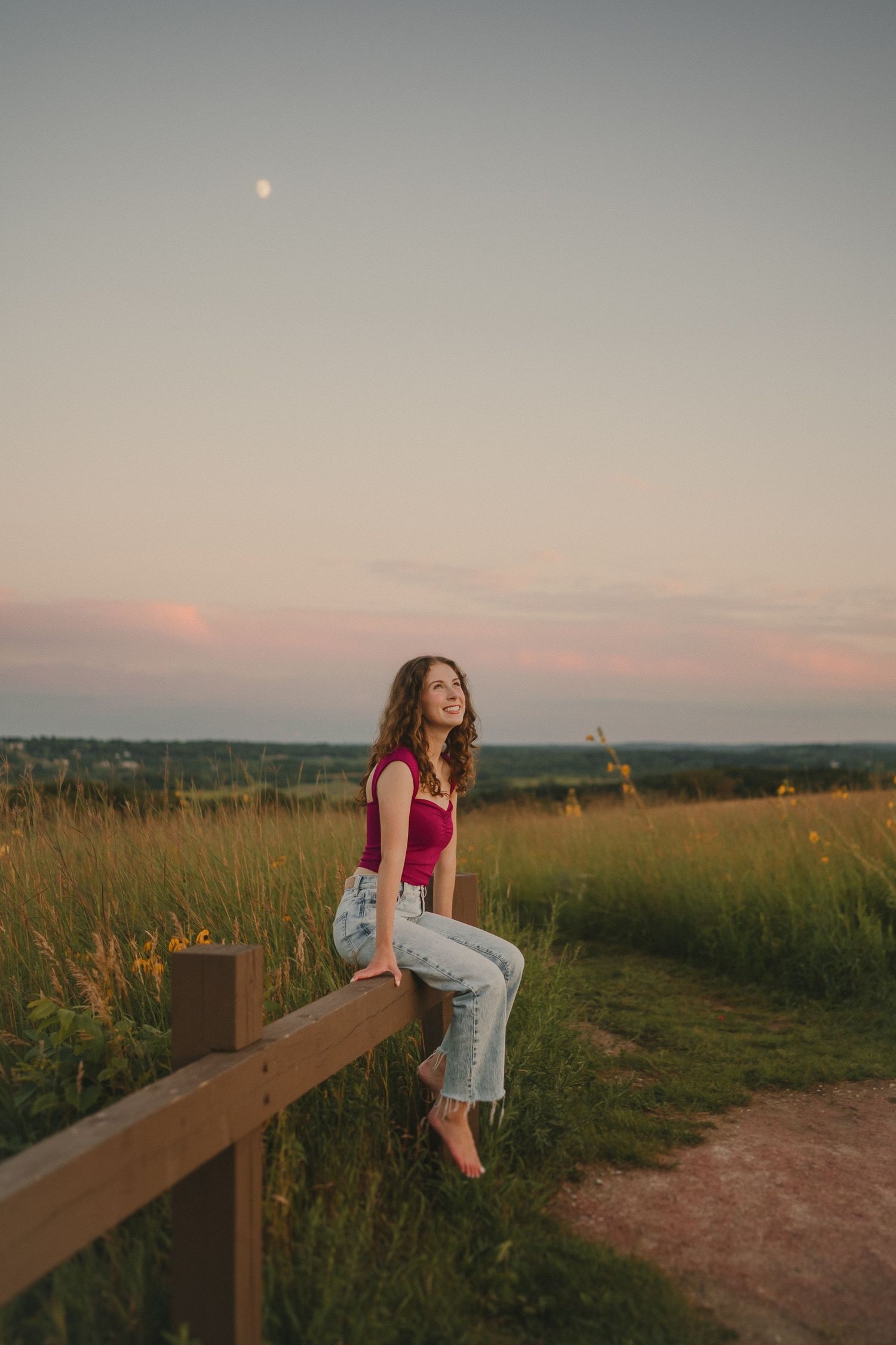 A casual-dressed person sits on a wooden fence overlooking a scenic prairie landscape at dusk.