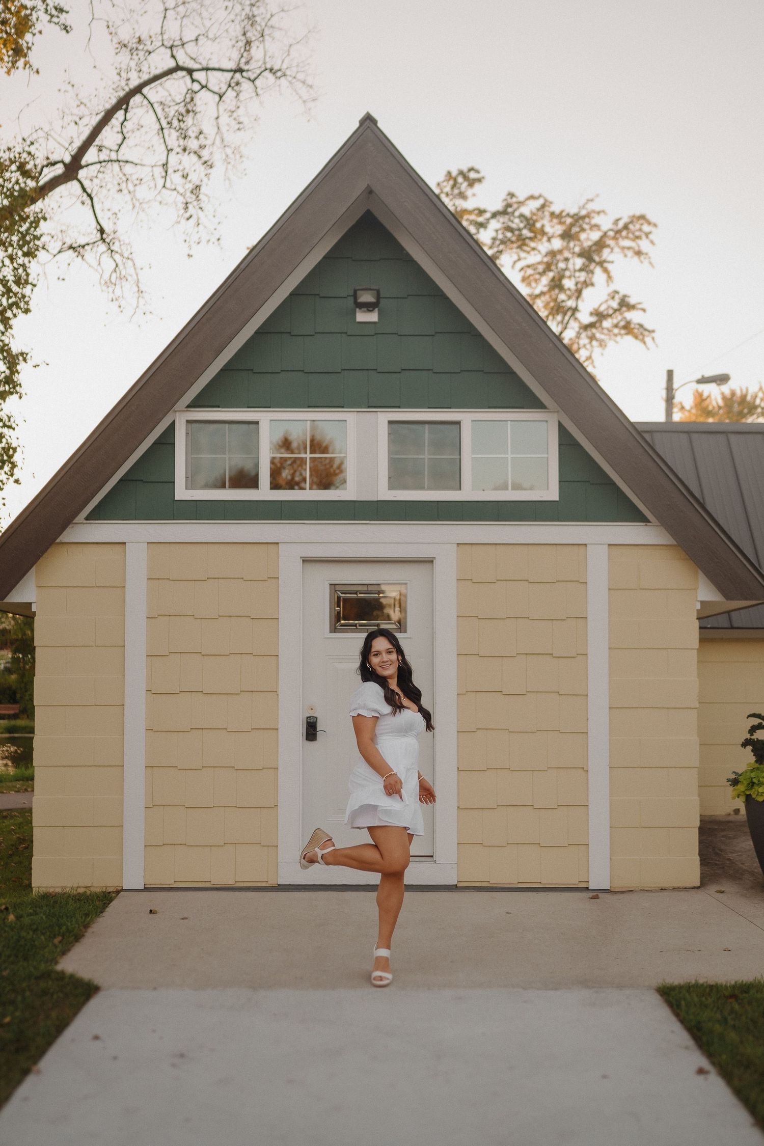 A person in a white dress poses playfully by the door of a small A-frame cottage with green trim.