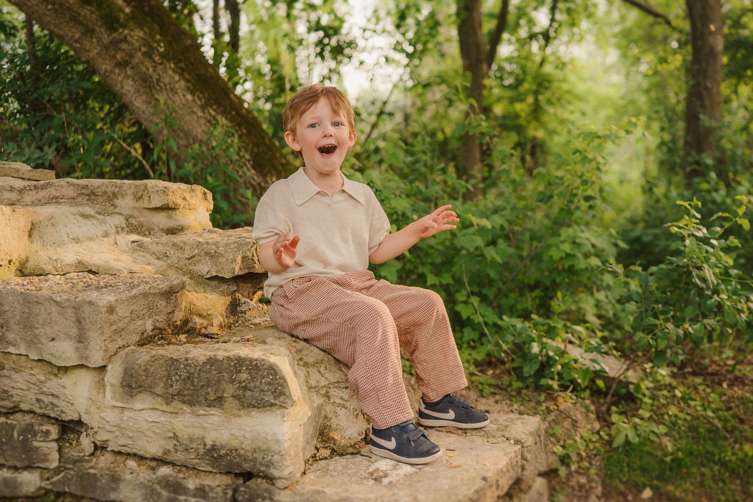 A child sits on stone steps outdoors, laughing with excitement against a green forest background.