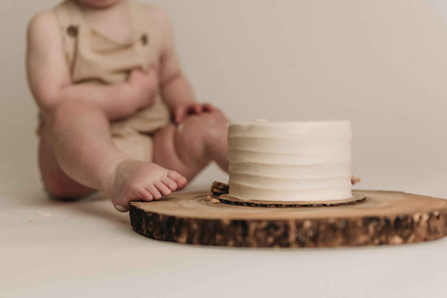 White cake on wooden slice with baby feet visible in soft focus background.
