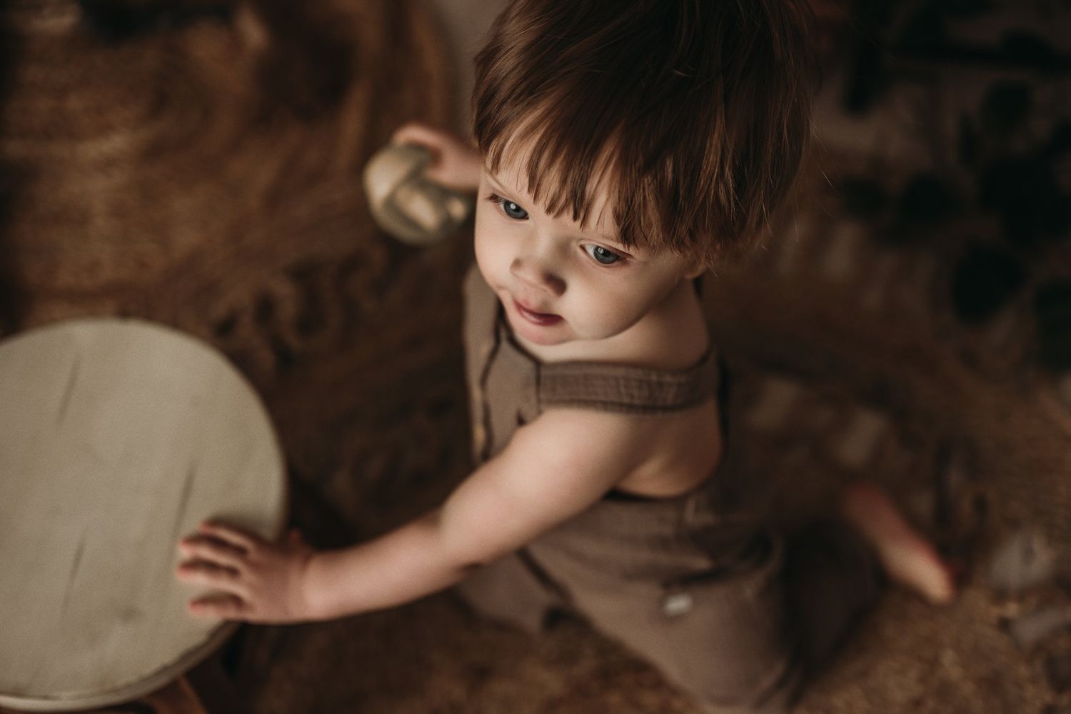 A toddler in a brown romper sits on a circular patterned rug in a cozy boho room.