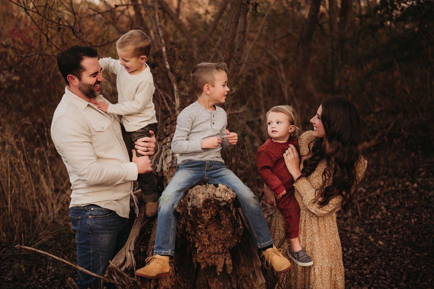A candid family moment captured in the woods with everyone gathered around a large tree stump during sunset.