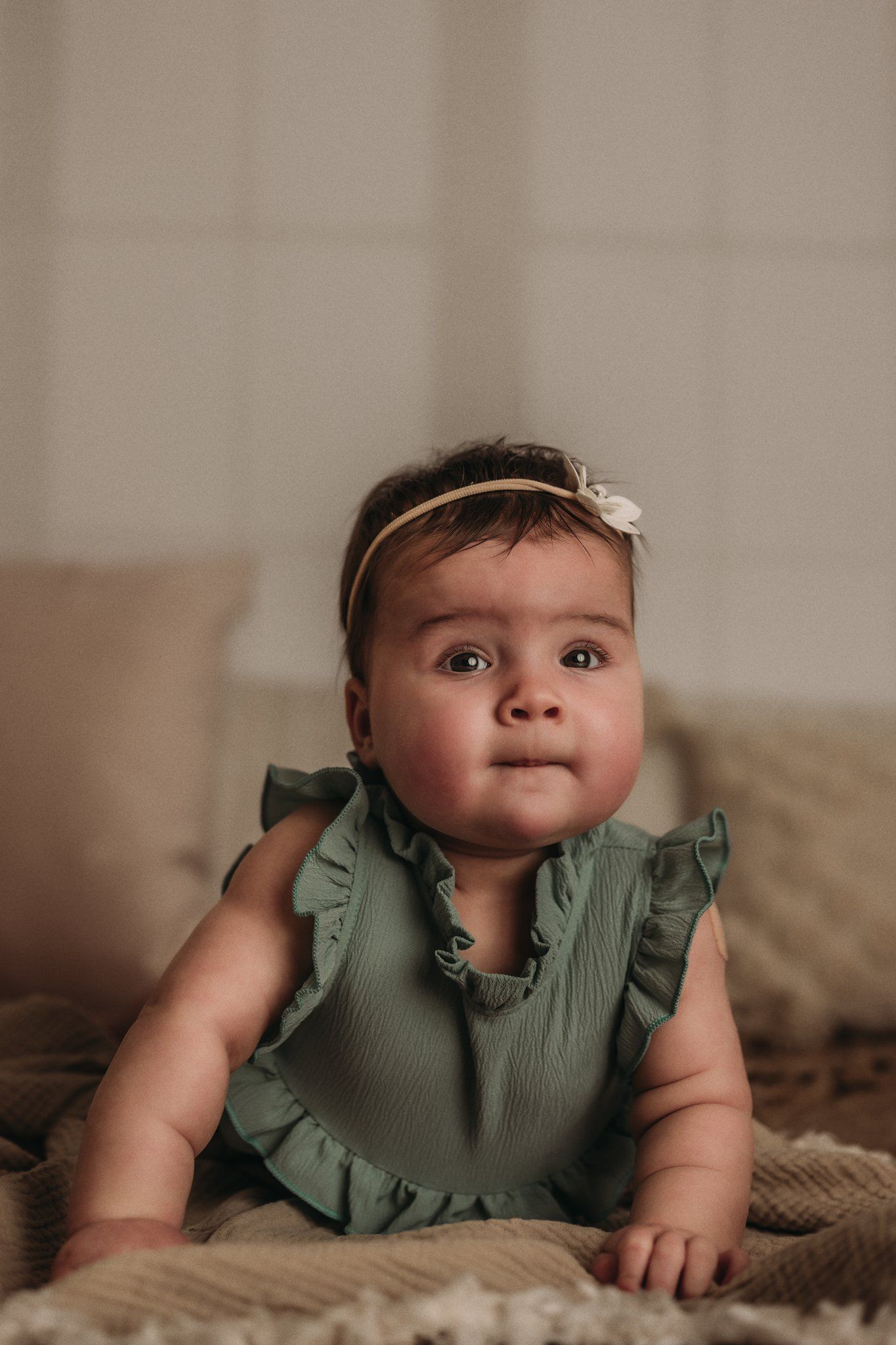 A baby in a mint green ruffled dress with headband poses on textured beige blankets.