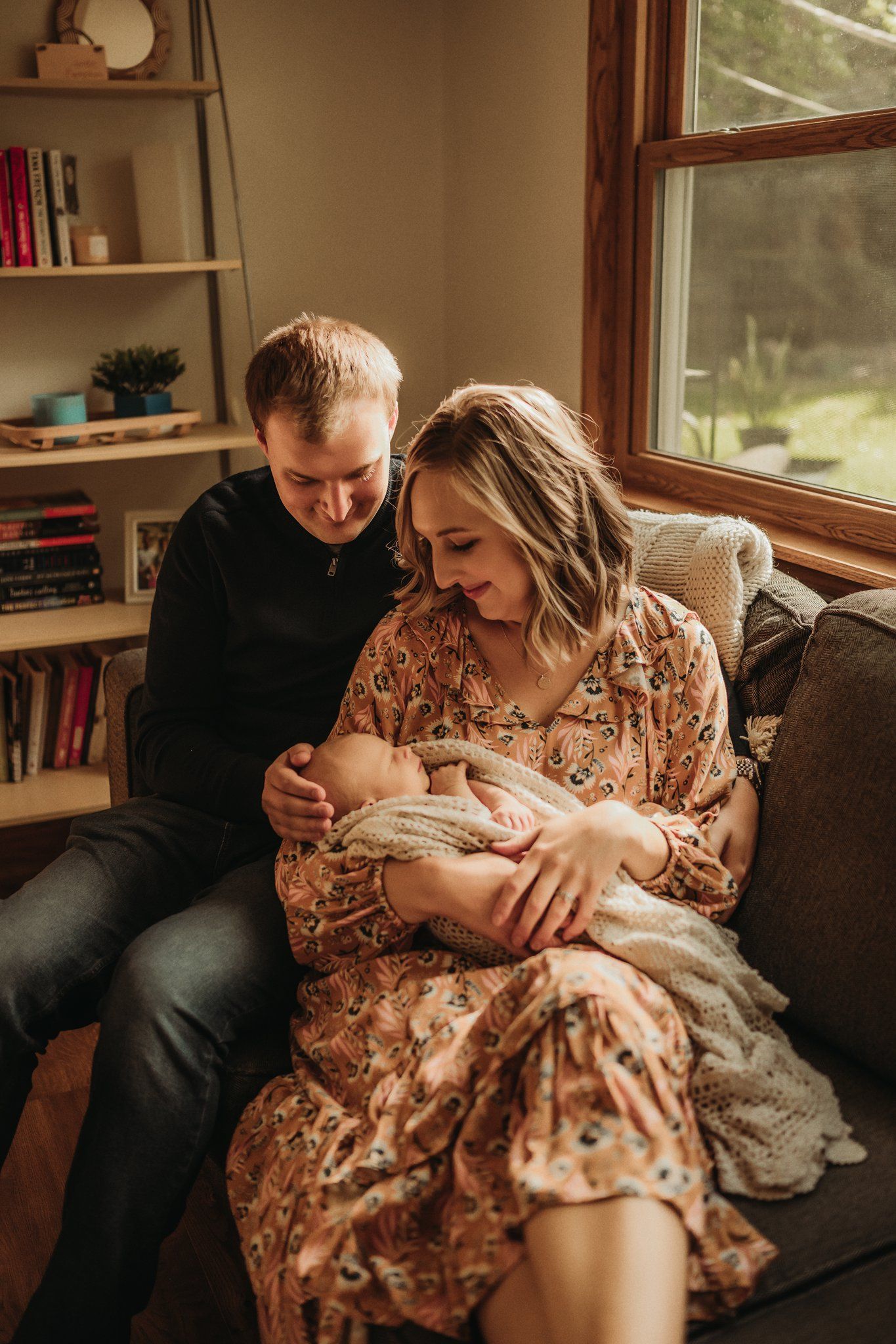Family lovingly cradles their newborn baby while sitting on a couch near a sunny window.