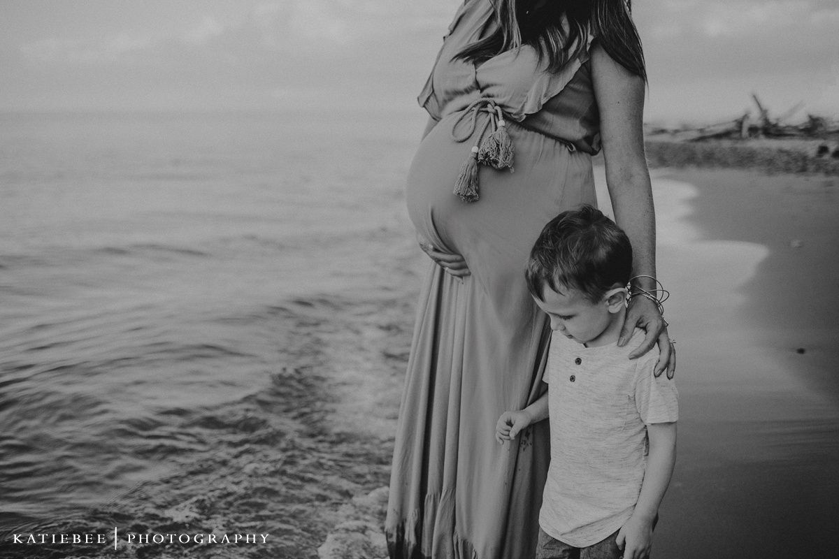 Black and white beach photo of pregnant woman and child looking at ocean.