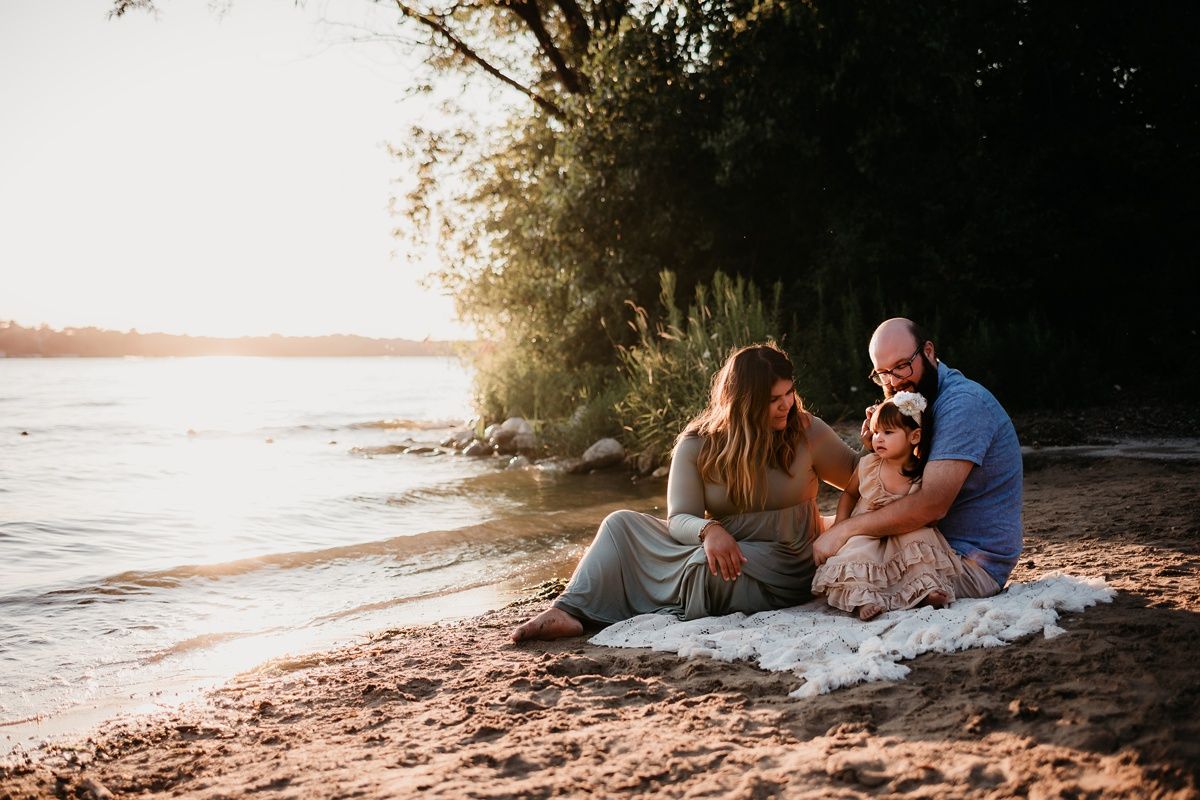 Family sitting on blanket at beach with sunset and trees in background.