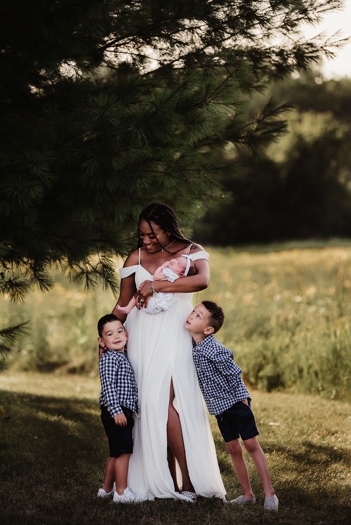 A touching outdoor family portrait showing a mother in a white dress embracing two children under a pine tree.