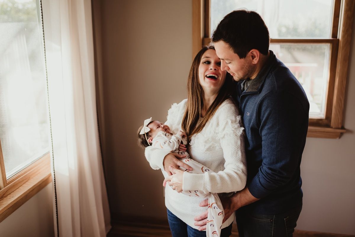 A tender moment between parents cradling their newborn baby near a sunlit window.