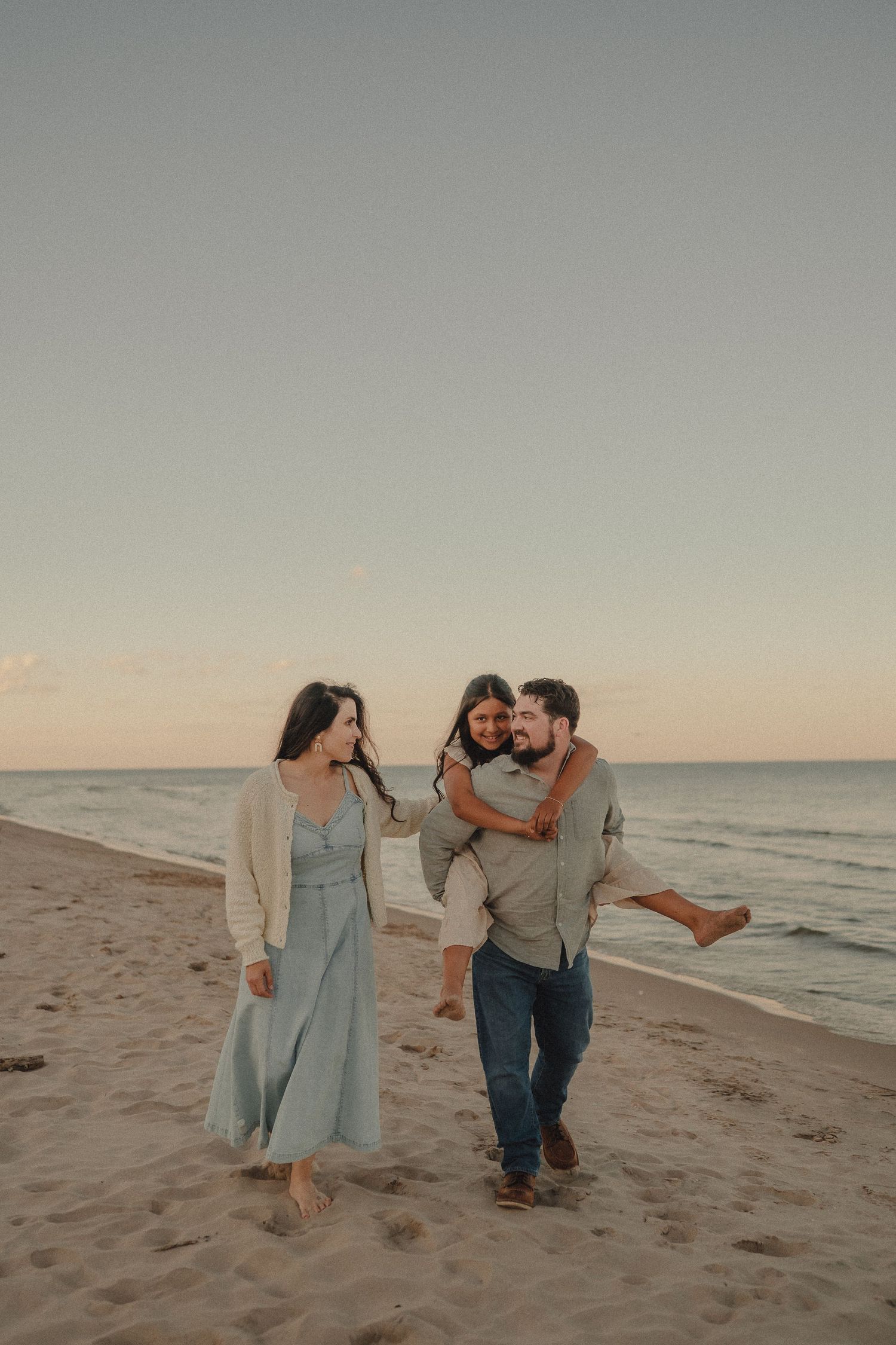 Family of three walking on a sandy beach at sunset, father giving daughter a piggyback ride.