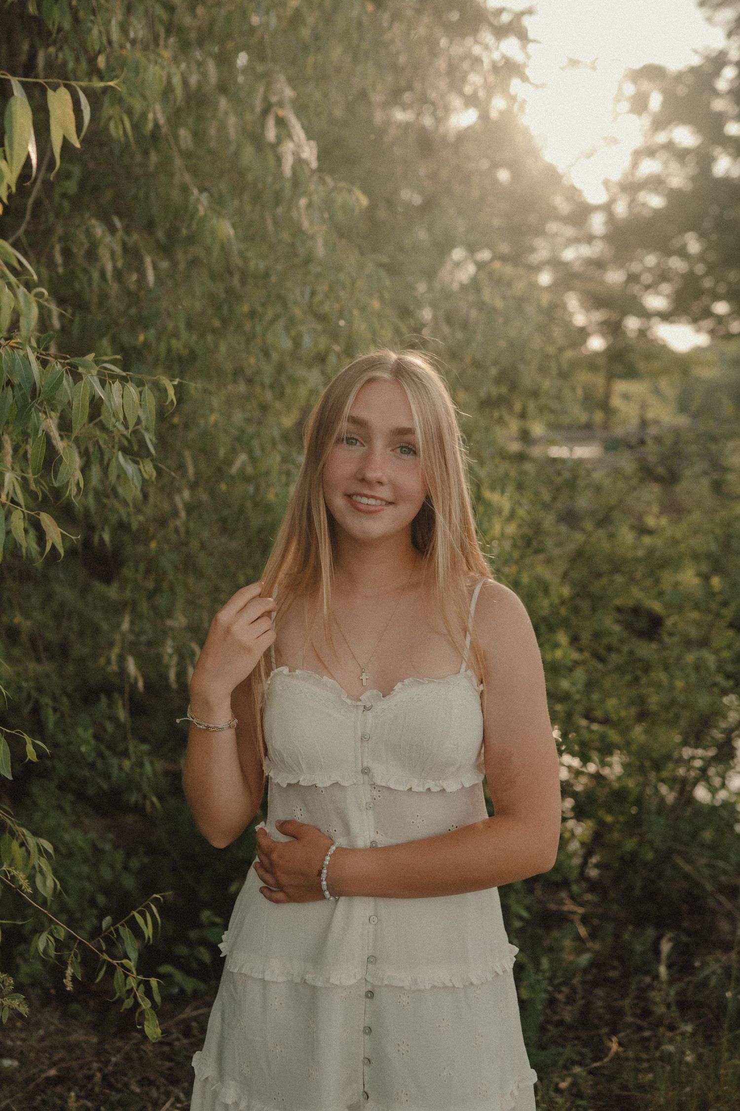 Young woman in white sundress smiling outdoors among lush green trees in golden hour sunlight.