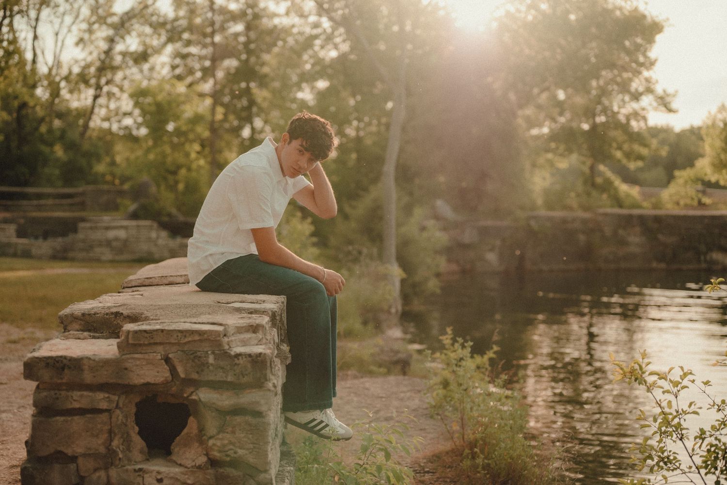 Young man sitting on stone wall by a pond, head in hands, contemplating in golden sunlight outdoors.